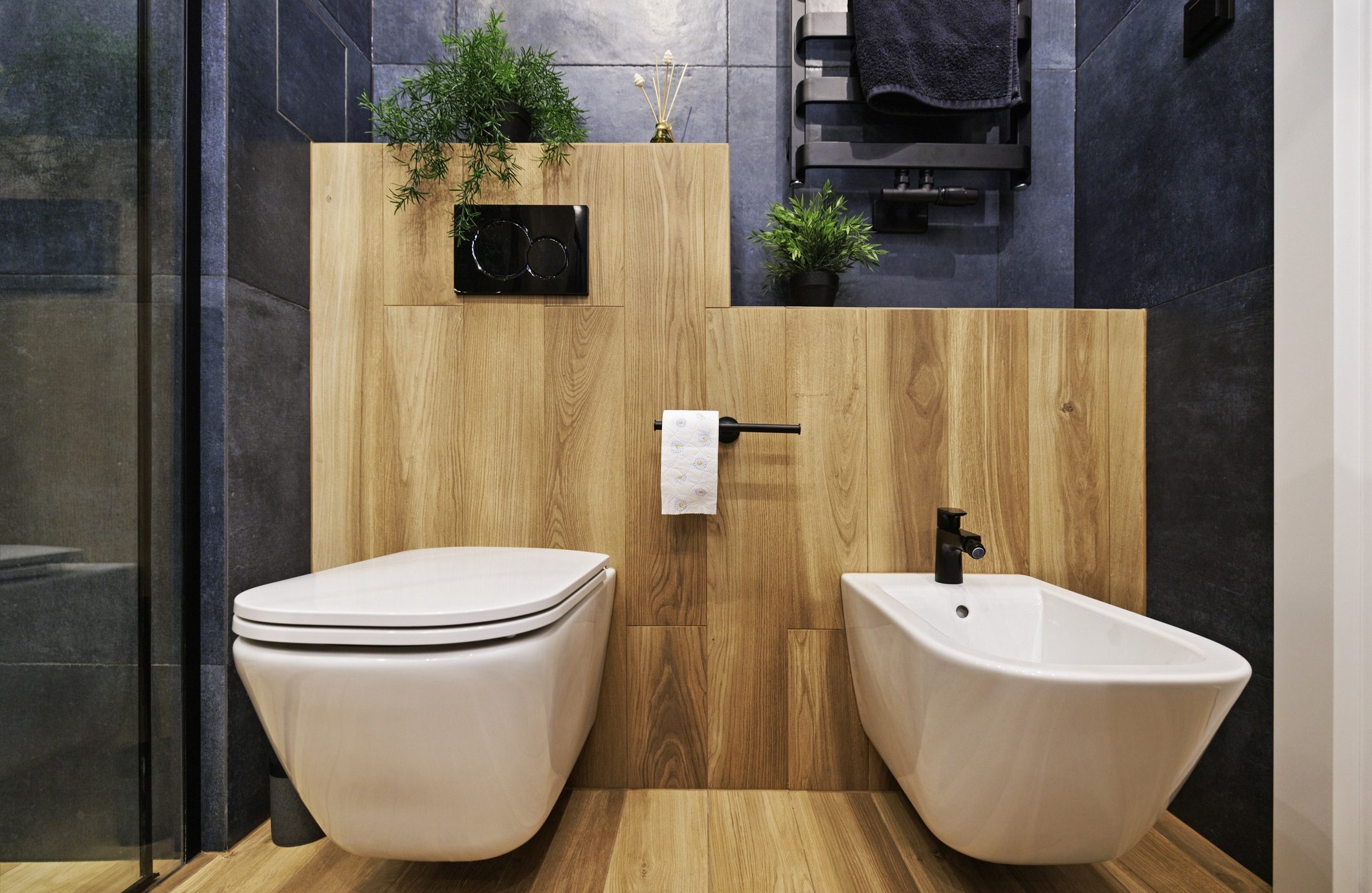 Modern bathroom featuring a wall-mounted toilet and bidet with white fixtures and wooden panels. A small black shelf holds two plants and a glass reed diffuser. Dark tiles surround the wood accents. Toilet paper holder and towel rack are also black.