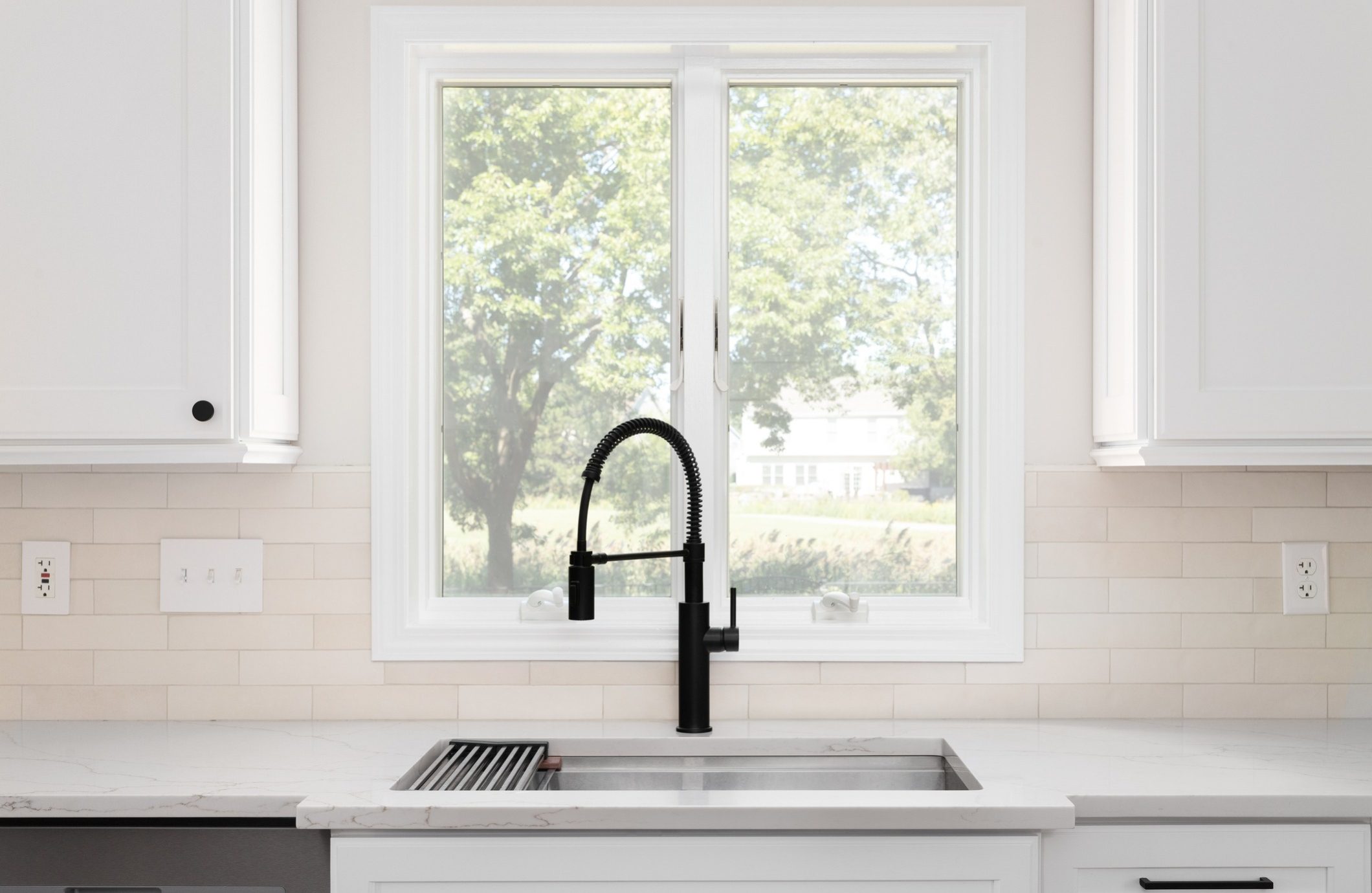 A modern kitchen sink with a black, flexible faucet is centered under a large, double-pane window. The countertop is white with a subtle marble pattern, and the backsplash features light beige subway tiles. Lush greenery is visible through the window.